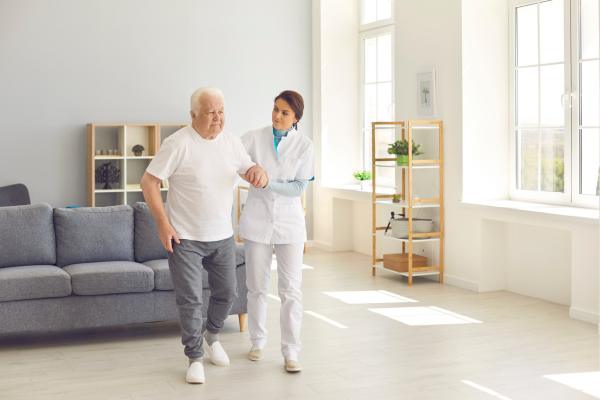 Un anciano caminando con la ayuda de una cuidadora en una residencia para mayores.